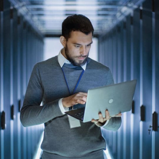 Man working on a laptop while standing in a corridor of servers